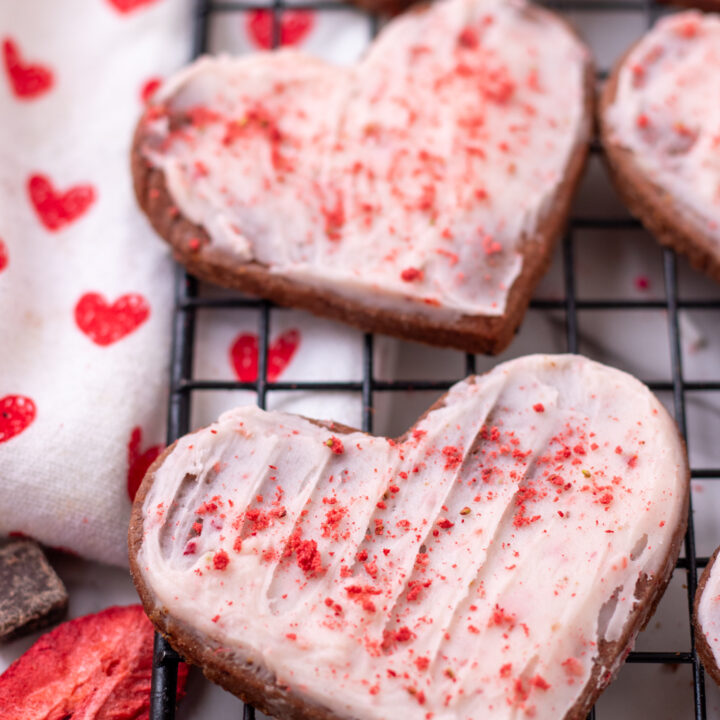A cooling rack topped with heart shaped chocolate sugar cookies with strawberry frosting. They're sprinkled with strawberry powder. There's a dish towel next to the cookies with little red hearts on it and a strawberry with chocolate chunks
