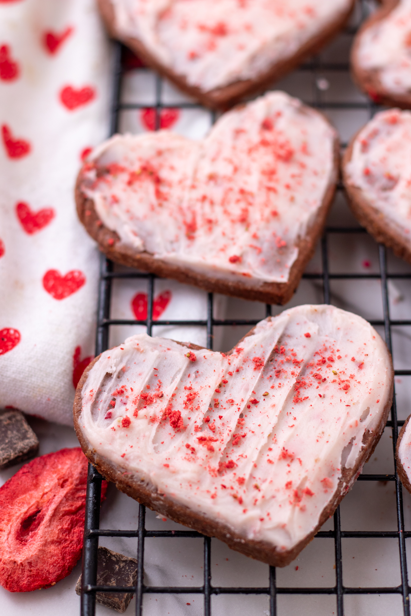 A cooling rack topped with heart shaped chocolate sugar cookies with strawberry frosting. They're sprinkled with strawberry powder. There's a dish towel next to the cookies with little red hearts on it and a strawberry with chocolate chunks
