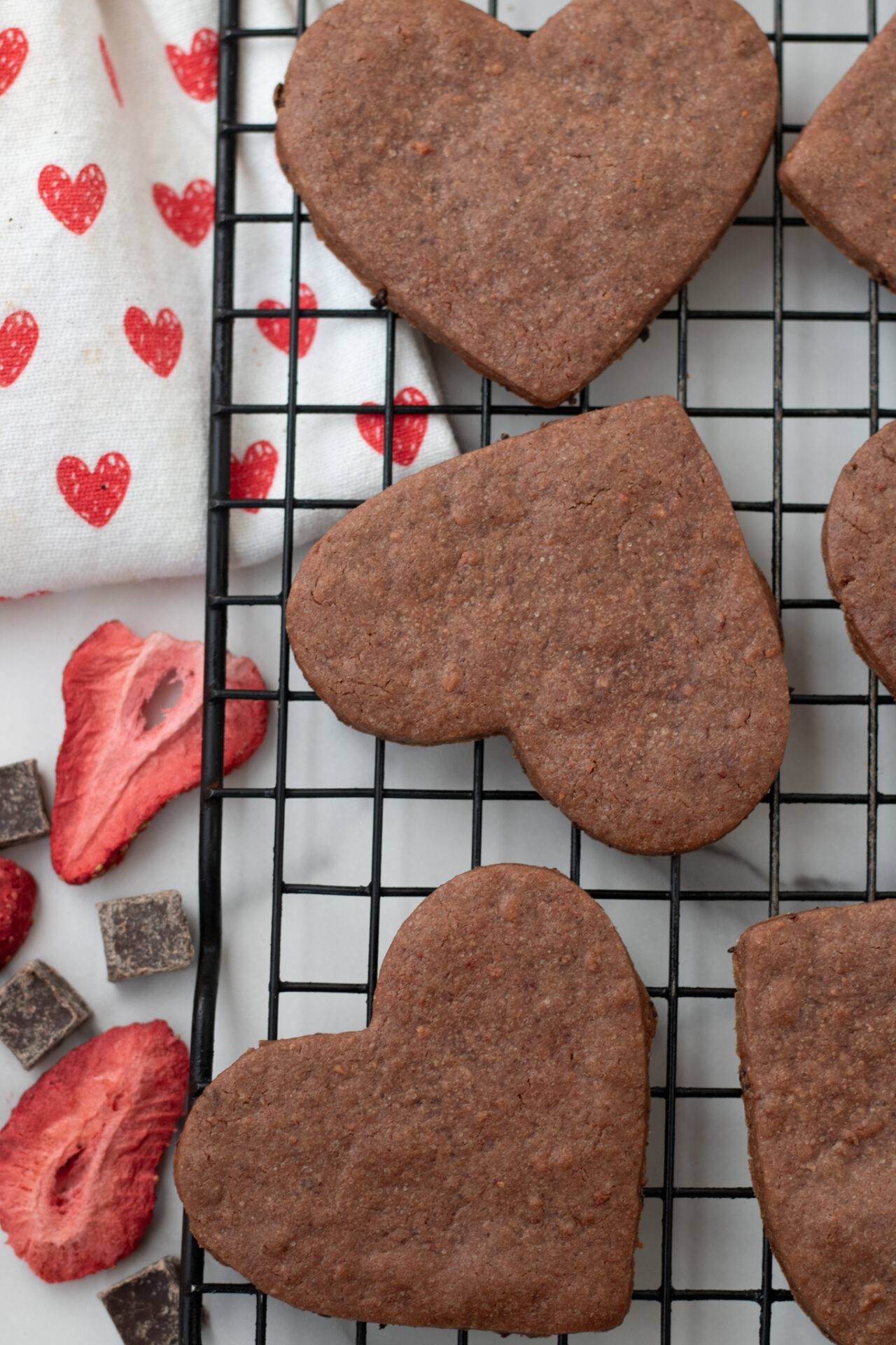 Heart shaped chocolate sugar cookies. There's a dish towel next to the chocolate cookies with little red hearts on it. There's a freeze dried strawberry and some chocolate chunks next to the cookies too.