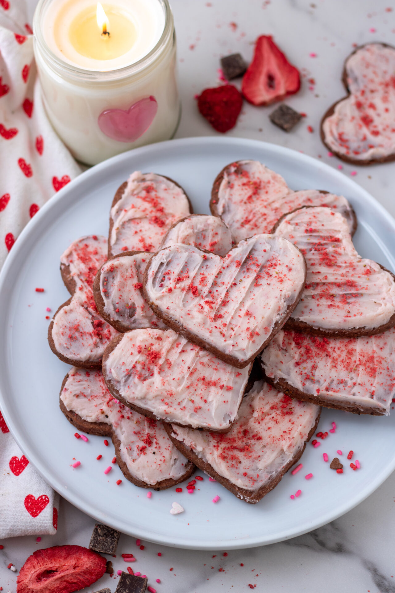 A white plate piled with heart shaped chocolate cookies with strawberry frosting. There's freeze dried strawberries and chocolate chunks in the background. A white candle is burning next to the plate of cookies.